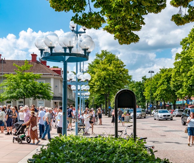 Livlig stadsgata med många människor, bilar, träd och gatlyktor en solig dag.