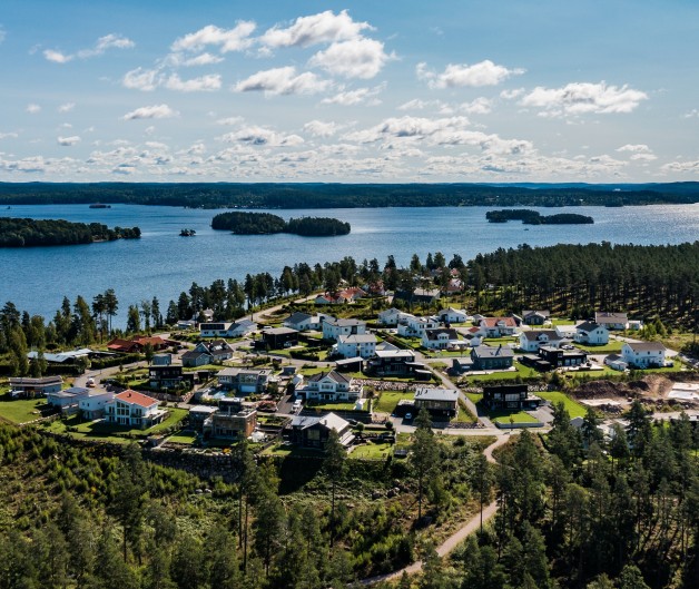 Flygbild över en liten by vid en sjö, omgiven av skog och vatten under en klarblå himmel.