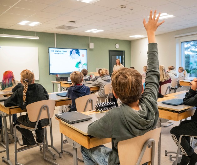 Ett klassrum med elever som sitter vid sina bänkar, en elev räcker upp handen och en lärare står framme vid tavlan.