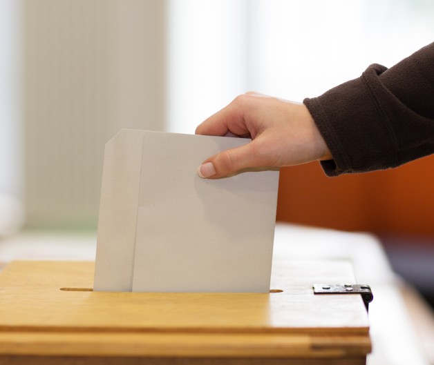 En hand lägger ett kuvert i en trälåda, troligen en valurna, inomhus.
