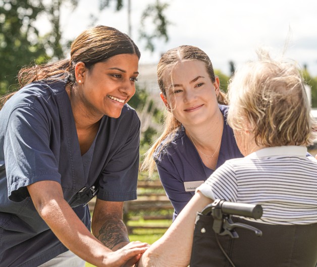 Två vårdare samtalar med en äldre person i rullstol utomhus en solig dag.