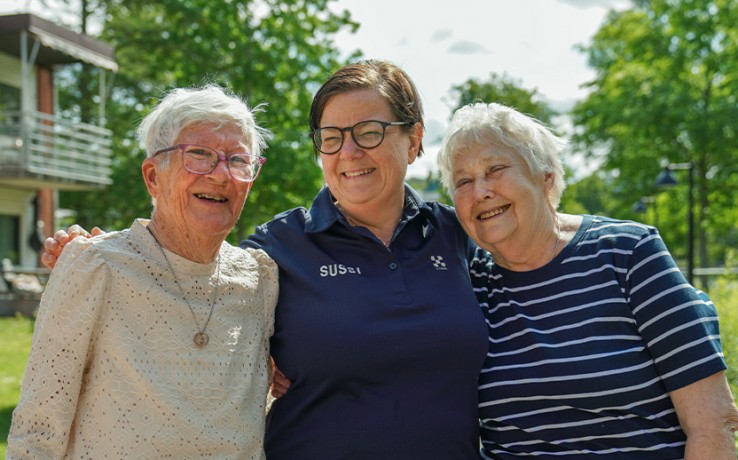 Tre personer står tillsammans utomhus, två äldre kvinnor och en yngre person i mitten, omgivna av grönska.