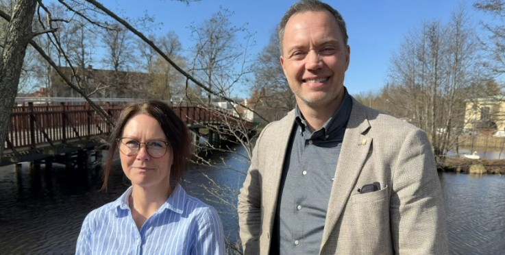 Två personer står utomhus vid en flod med en träbro och träd i bakgrunden, ansikten är suddade.